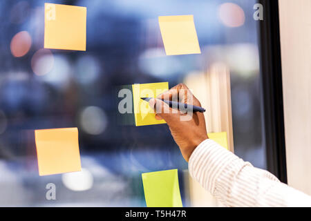 La mano di imprenditore scrivere con la penna sulla nota adesiva incollaggio sul vetro di finestra, close-up Foto Stock