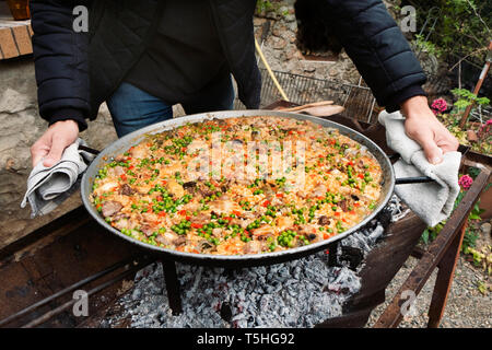 Primo piano di un giovane uomo caucasico con una tipica paella spagnola, cotto in una padella per paella con fuoco di legna, all'aperto Foto Stock