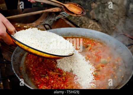 Primo piano di un giovane uomo caucasico preparare una tipica paella spagnola, in una padella per paella con fuoco di legna, all'aperto Foto Stock