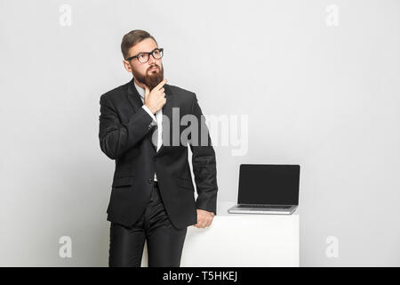 Grave fiducioso pensieroso barbuto giovane imprenditore in abito nero sono in piedi vicino al suo luogo di lavoro e tenendo la sua barba con faccia di concentrazione Foto Stock