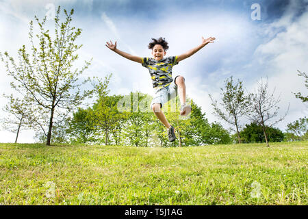 Felice energico giovane ragazzo saltare in aria in alto come lui corre attraverso l'erba in un parco o giardino con le braccia tese in un basso angolo di visione Foto Stock