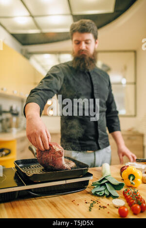 Barbuto chef cucinare la carne in una padella in cucina. L'uomo la preparazione di Bollito di maiale sulla tavola fornelli elettrici Foto Stock