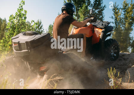 Atv rider salire la montagna di sabbia nella cava, vista posteriore, nubi di polvere. Driver maschio nel casco su quad bike, offroad nella buca di sabbia Foto Stock