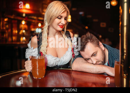 Bella cameriera con boccale di birra si affaccia su uomo ubriaco al bancone in pub, oktoberfest vacanze. Persona di sesso maschile di bere in bar, barista in tradizionale retr Foto Stock