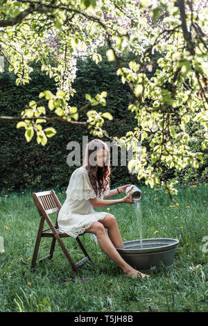 Giovane donna indossa abito bianco Preparazione pediluvio in giardino Foto Stock