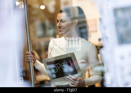 Donna matura in piedi in shop , sorridente, con open firmare appeso nella finestra Foto Stock