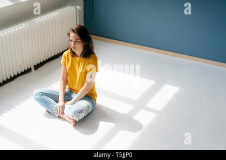 Malinconici donna seduta in presenza di luce solare a gambe incrociate sul pavimento Foto Stock