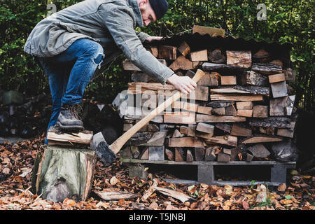 Uomo Barbuto vestita di nero beanie e parka in piedi nel giardino in autunno, utilizzando ax per tritare un pezzo di legno sul blocco di trinciatura. Foto Stock