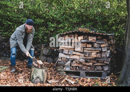 Uomo Barbuto vestita di nero beanie e parka in piedi nel giardino in autunno, utilizzando ax per tritare un pezzo di legno sul blocco di trinciatura. Foto Stock