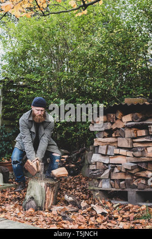 Uomo Barbuto vestita di nero beanie e parka in piedi nel giardino in autunno, utilizzando ax per tritare un pezzo di legno sul blocco di trinciatura. Foto Stock