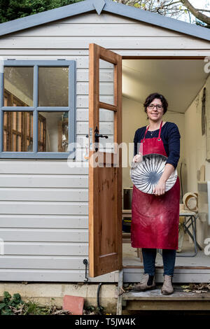 Donna che indossa il grembiule rosso in piedi sui gradini fuori la sua officina, tenendo la coppa in ceramica con linea nera pattern. Foto Stock