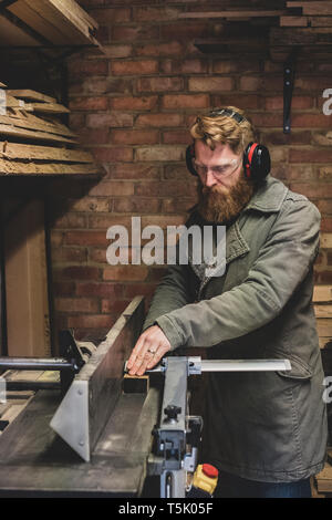 Uomo Barbuto in piedi in officina, indossare protezioni per le orecchie, lavorando sul pezzo di legno. Foto Stock