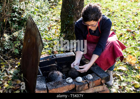 Angolo alto close up della donna che indossa il grembiule rosso inginocchiato accanto al fumo outdoor fire pit, lavorando sul vaso. Foto Stock