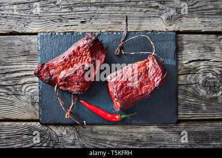 Due pezzi di barbecue affumicati il filetto di carne con spago su un vassoio di pietra su un vecchio tavolo di legno, vista da sopra, laici piatta Foto Stock