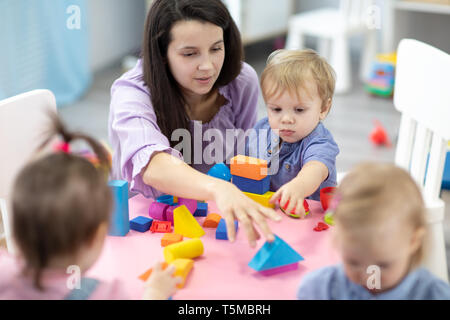 Insegnante femmina seduti al tavolo nella sala da gioco con tre bambini asilo costruzione Foto Stock