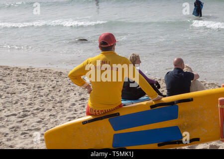 Bagnino RNLI sat sulla tavola da surf in un affollato Sennen Cove Beach, Cornwall Foto Stock