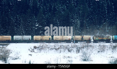Treno merci si muove attraverso la foresta di conifere durante la nevicata lungo il fiume congelato tra colline. Nord del paese. Lato fron top Foto Stock