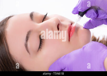 Bellissima fanciulla getting tatuaggio labbra rosse in studio di bellezza. Professional cosmetologo rendendo trucco permanente con utensile in viola i guanti Foto Stock