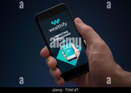 Un uomo guarda al suo iPhone che visualizza il logo Wealthify (solo uso editoriale). Foto Stock