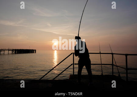 Pesca sportiva fisherman mediante aste di spiaggia espressi dal marciapiede al di sopra Totland Bay come il sole tramonta oltre il lontano orizzonte e Totland Pier. Foto Stock