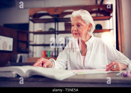 Elegante anziana signora con le rughe del viso seduta a tavola e libro di lettura Foto Stock