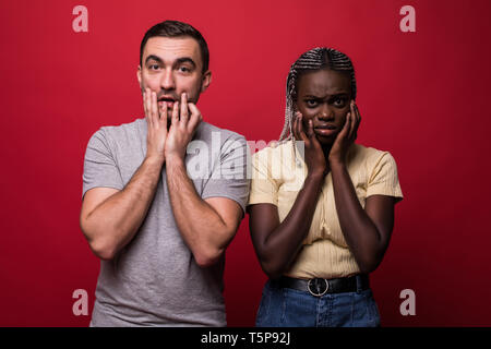 Ritratto di scioccato interracial giovane: donna africana e uomo caucasico in piedi insieme contro la parete rossa, che ricopre la faccia, guardando la telecamera con Foto Stock