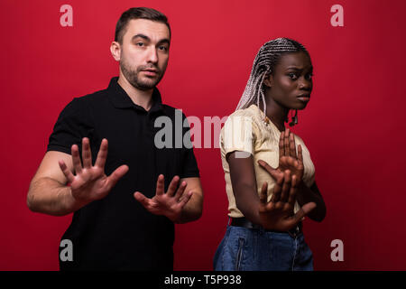 Ritratto di scioccato interracial giovane: donna africana e uomo caucasico in piedi insieme contro la parete rossa, che ricopre la faccia, guardando la telecamera con Foto Stock