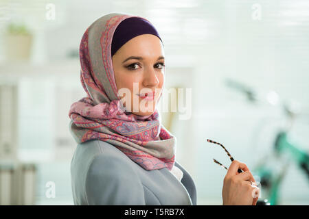 Bella donna elegante in hijab e occhiali, seduti alla scrivania con computer portatile in ufficio. Ritratto di fiducioso imprenditrice musulmana. Ufficio moderno con Foto Stock