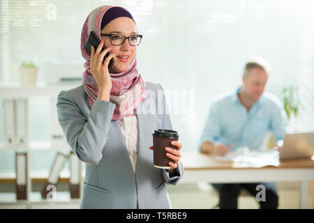 Bella donna di hijab e occhiali parlando al telefono, sorridente, tenendo tazza di caffè. Ritratto di fiducioso imprenditrice musulmana. Ufficio moderno Foto Stock