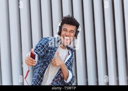 Giovane mulatto uomo che indossa le cuffie in piedi isolato su una strada di città ascoltando la musica puntando alla fotocamera a sorridere giocoso che mostra il pollice in alto Foto Stock