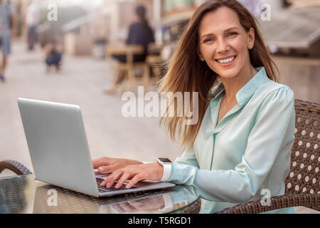 Giovane donna seduta al tavolo nell'urbano cafe lavorando su laptop cercando fotocamera a sorridere gioiosa ventoso Foto Stock