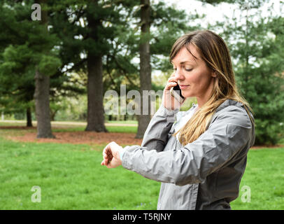 Giovane donna sulla cella, telefono mobile guardando a guardare nel parco della città su nuvoloso giorno, sorridente, con l'erba verde, alberi sullo sfondo Foto Stock