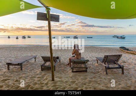 Bella donna di fotografare il tramonto sulla spiaggia idilliaca seduta con il mare e le barche in background. La pace e la consapevolezza del concetto. Bahia, Brasile. Foto Stock