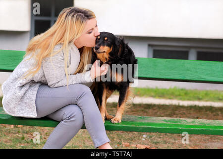 Bella donna con il suo piccolo di razza dog sitter e in posa nella parte anteriore della fotocamera sulla panca in legno presso il parco della città. Ritratto del proprietario e la cute cane. Foto Stock