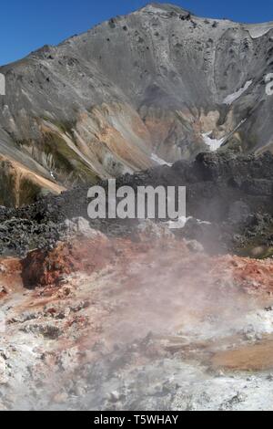 Vista sulla vallata con colorate di rosso, bianco e nero pietre fumante con robusto grigio faccia di montagna in contrasto con il cielo blu senza nuvole - Landmannalaugar, Foto Stock