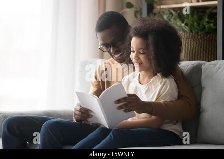 Padre nero libro di lettura alla figlia seduta sul lettino Foto Stock