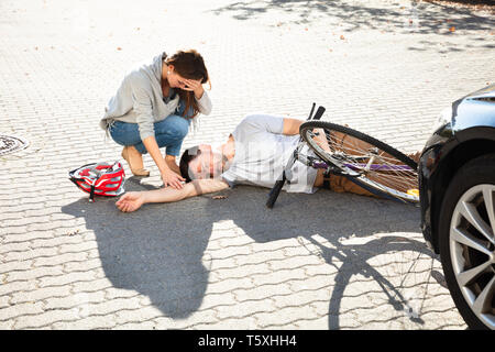 Giovane donna Guardando inconscio ciclista maschio giacente sulla strada dopo un incidente nei pressi di auto Foto Stock