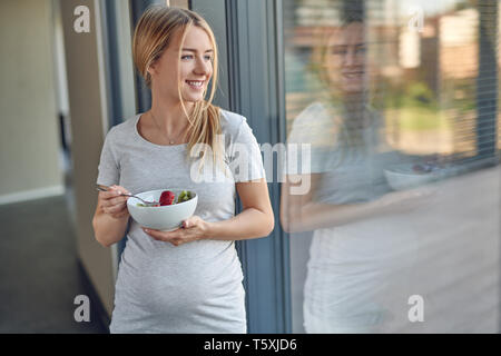 Felice gravide sane bionda e giovane donna in piedi appoggiata contro una finestra patio sorridente come lei si guarda all'aperto godendo di una coppa di frutta fresca sa Foto Stock