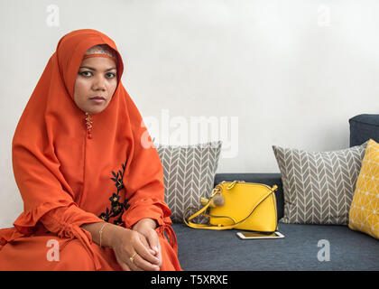 Donna asiatica indossando orange hijab outfit seduti sul divano guardando la fotocamera. Foto Stock