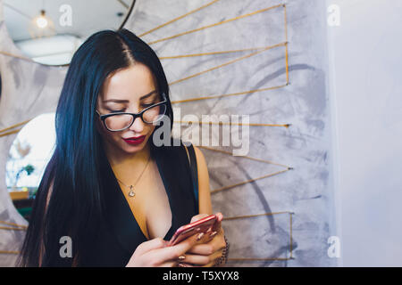 Felice ragazza araba utilizzando smart phone su un muro di mattoni. Donna sorridente con capelli ricci in abiti casual in background urbano. Foto Stock