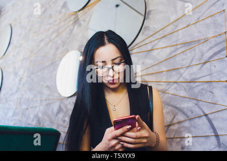 Felice ragazza araba utilizzando smart phone su un muro di mattoni. Donna sorridente con capelli ricci in abiti casual in background urbano. Foto Stock