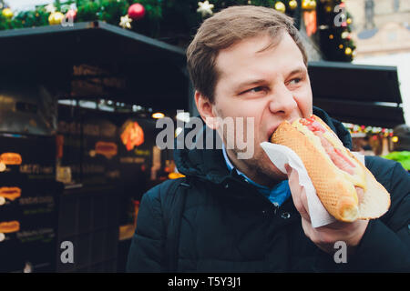 Closeup ritratto di fame uomo in bicchieri di mangiare hot dog a sfondo all'aperto. Foto Stock