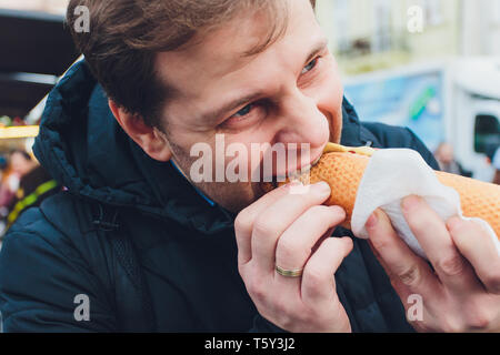 Closeup ritratto di fame uomo in bicchieri di mangiare hot dog a sfondo all'aperto. Foto Stock