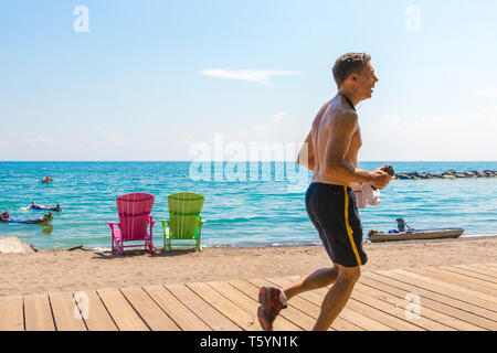 Uomo anziano in esecuzione passato due luminose colorate deck o sdraio in spiaggia con la chiave balsamico beach affacciato sul lago Ontario in East Toronto, Canada Foto Stock