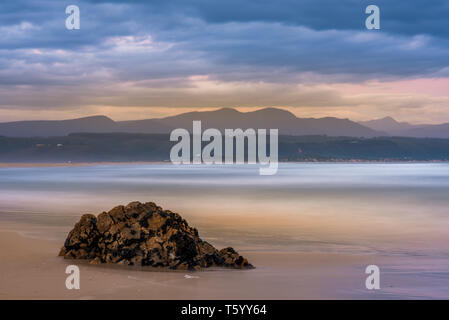 Trasognata di onde che si infrangono in slow motion a Plettenberg Bay spiaggia al tramonto, con colline in lontananza e una roccia in primo piano. Garden Route, Wester Foto Stock