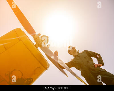 Un bel giovane pilota in un verde nel complesso in piedi accanto al propulsore di un vecchio piano in una giornata di sole. Foto Stock