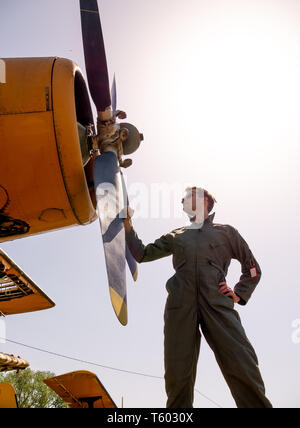 Un bel giovane pilota in un verde nel complesso in piedi accanto al propulsore di un vecchio piano in una giornata di sole. Foto Stock