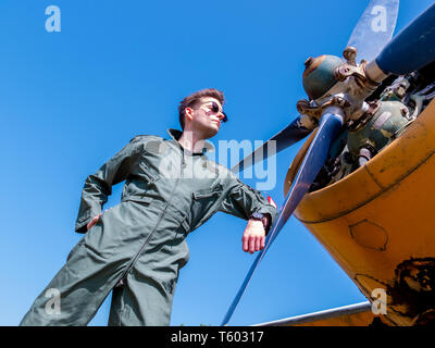 Un bel giovane pilota in un verde nel complesso in piedi accanto al propulsore di un vecchio piano in una giornata di sole. Foto Stock