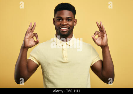 Guy positivo in elegante T-shirt mostra OK gesto con entrambe le mani su uno sfondo giallo., dimostrando che tutto è andato bene, mostrando la sua approvazione, Foto Stock