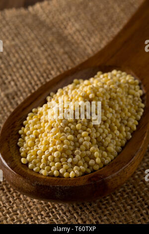 Pila di miglio dorato, un senza glutine di sementi di grano, in cucchiaio di legno su tela di tessuto su brown cucina rustica tavolo - messa a fuoco selettiva Foto Stock